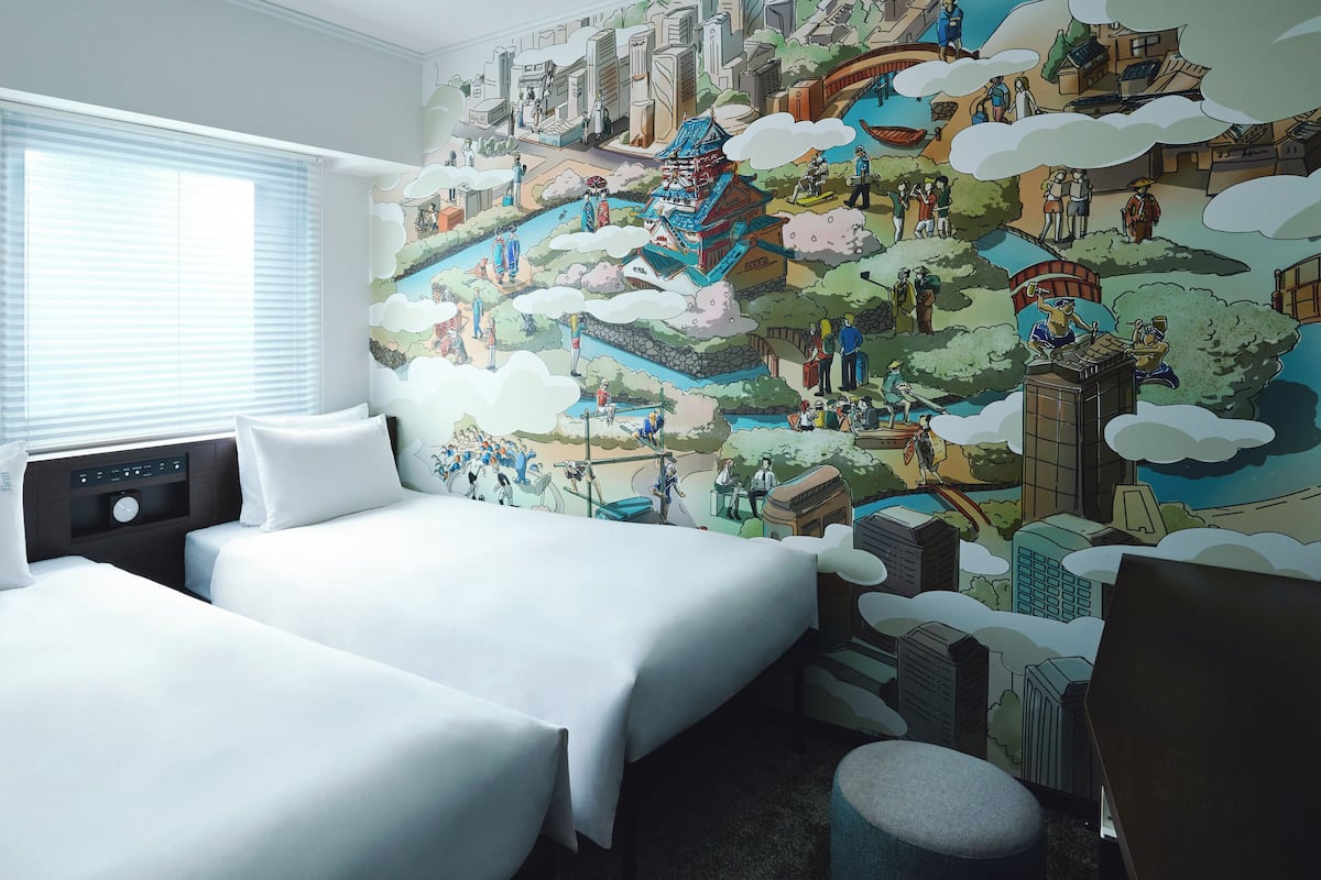 Une chambre d’hôtel avec deux lits, une fenêtre avec des stores, une fresque murale et un petit pouf rond.