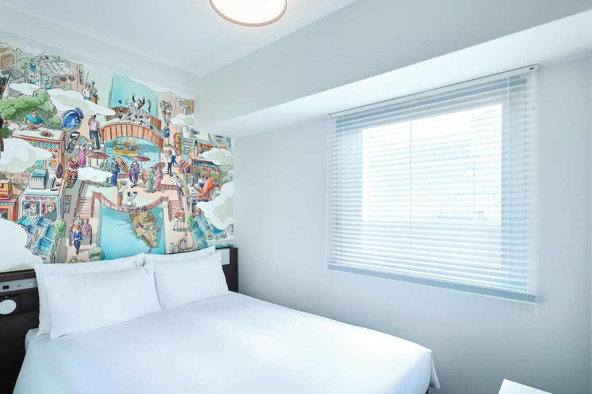 Une chambre d’hôtel avec un grand lit, une table de chevet, une commode, une fenêtre avec des stores et une fresque colorée sur le mur.