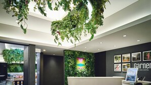 Un espace d’accueil d’hôtel au design moderne, comprenant un comptoir d’accueil, un mur orné de tableaux et un plafond décoré de plantes suspendues.