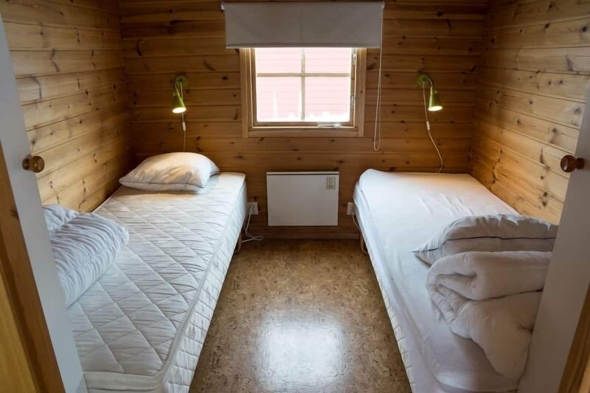 Una pequeña habitación de cabaña de madera con dos camas, una ventana y un calentador fijado a la pared.