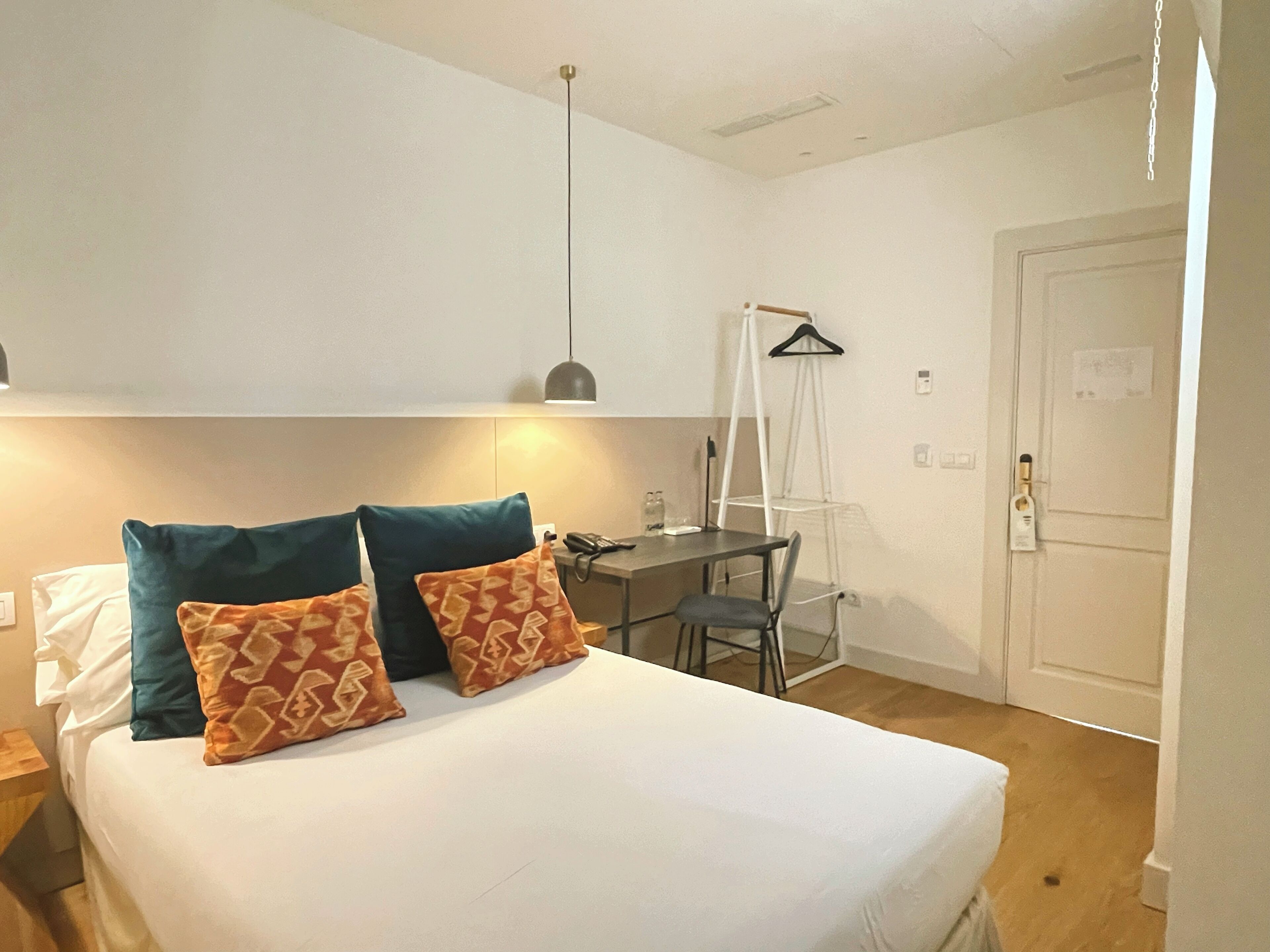 Ein Schlafzimmer mit Bett, Kissen, Schreibtisch, Stuhl, Pendelleuchte und einer Tür.