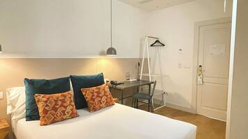 A bedroom with a bed, pillows, a desk, a chair, a hanging light, and a door.
