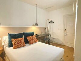A bedroom with a bed, pillows, a desk, a chair, a hanging light, and a door.