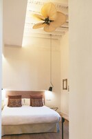 Un dormitorio con una cama, dos almohadas estampadas, un ventilador de techo y una mesita de noche con una lámpara.