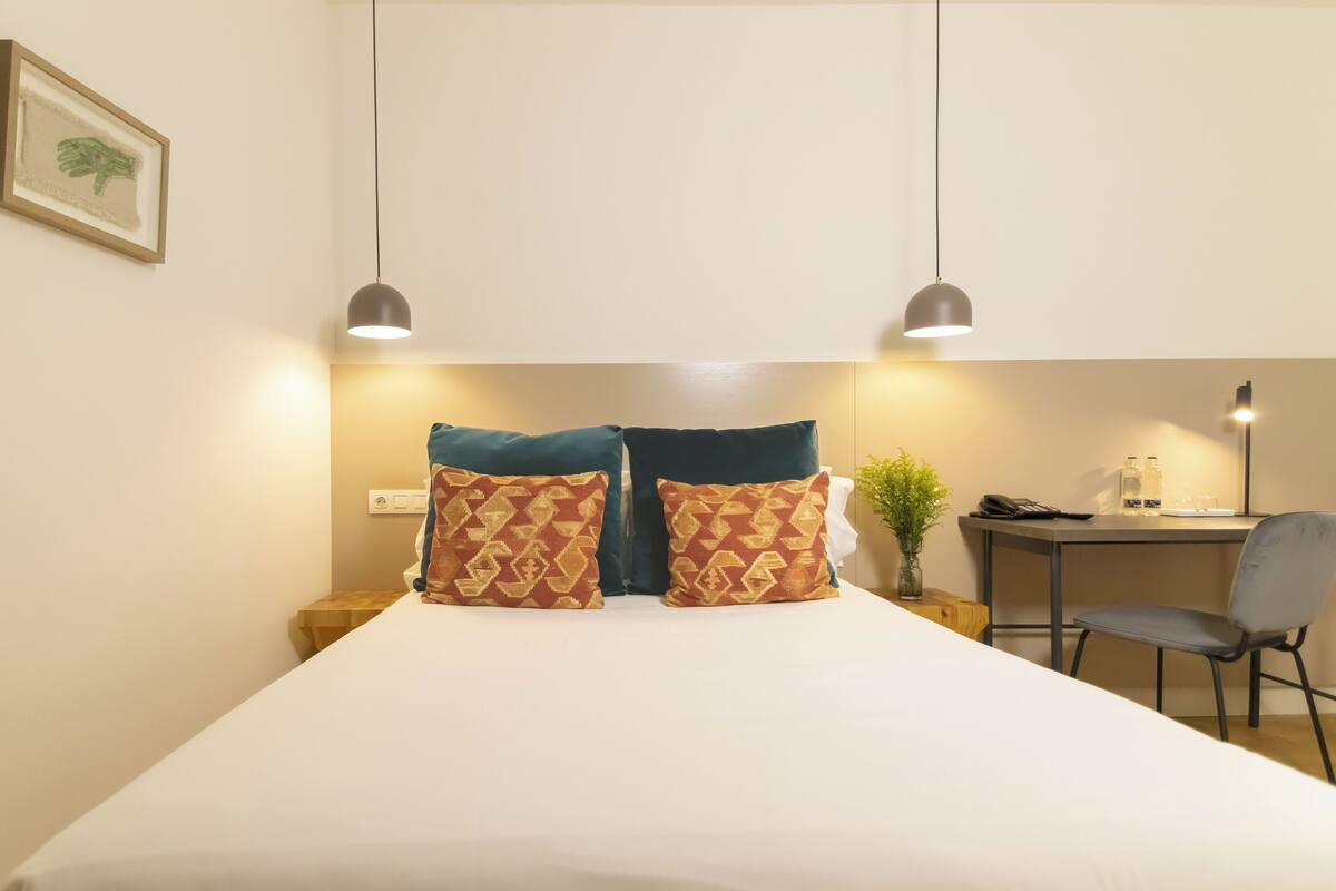A hotel room with a large bed, two patterned pillows, a desk with a chair, and a small table with a plant.