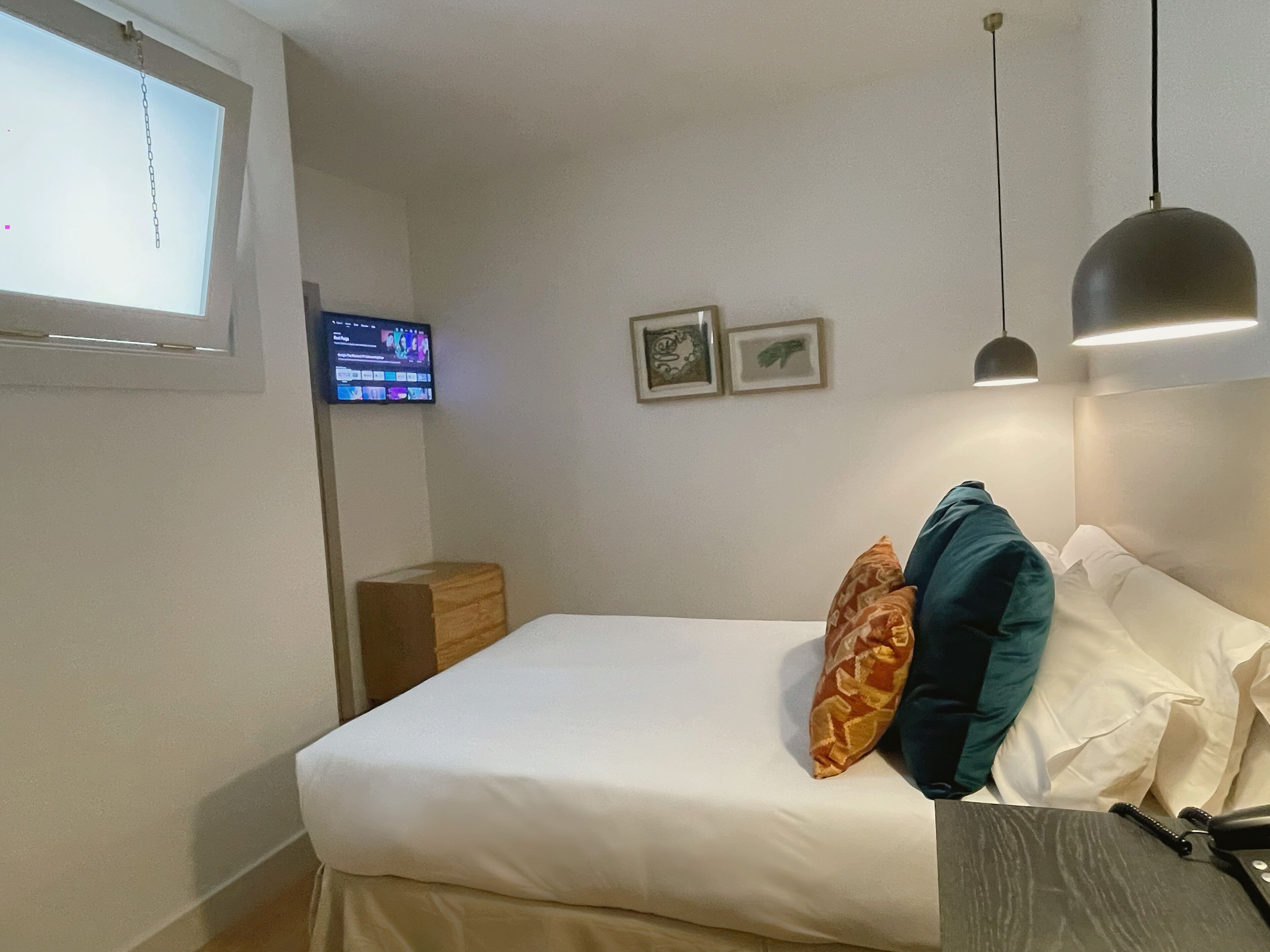 A hotel room with a bed, a desk, a chair, a lamp, a TV, and two framed pictures on the wall.