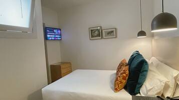 A hotel room with a bed, a desk, a chair, a lamp, a TV, and two framed pictures on the wall.