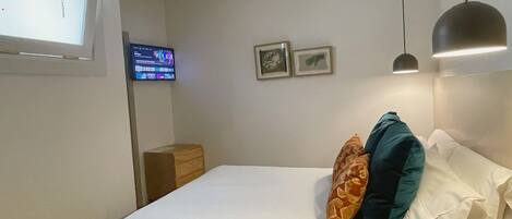 A hotel room with a bed, a desk, a chair, a lamp, a TV, and two framed pictures on the wall.