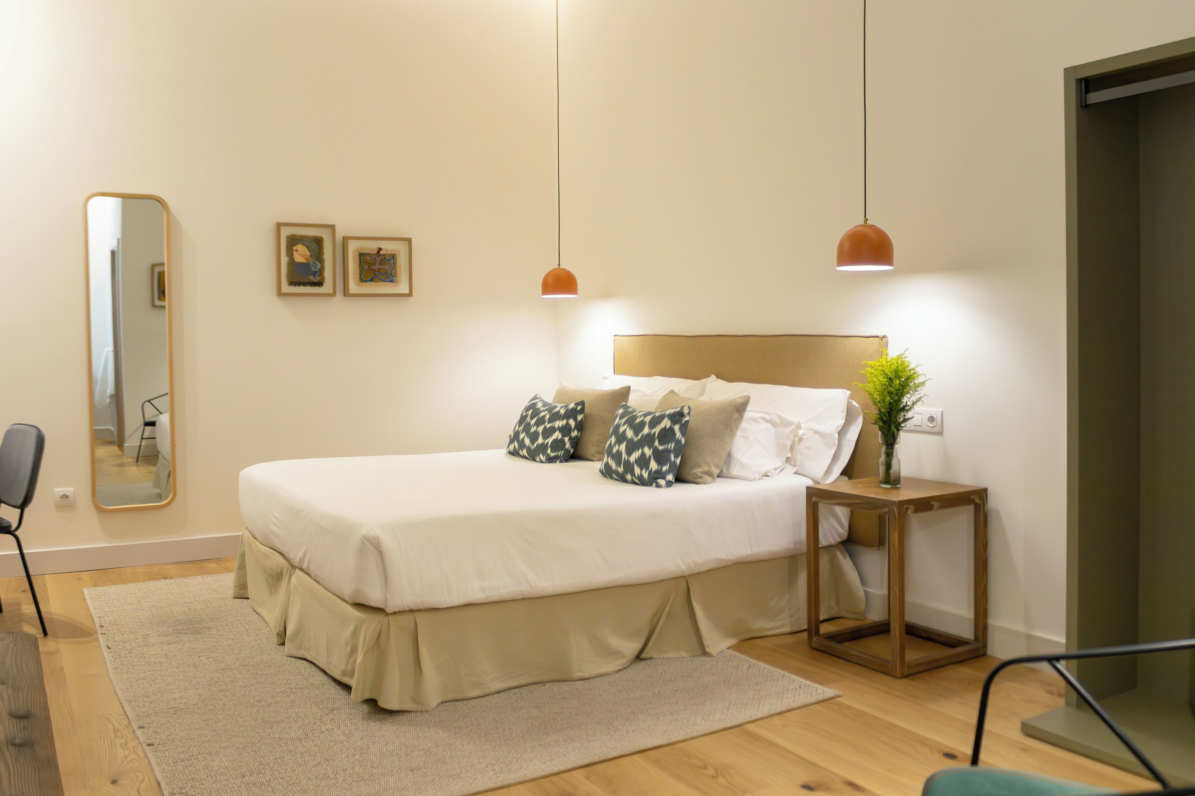 Un dormitorio ordenado con cama, mesita de noche, espejo y una planta pequeña.