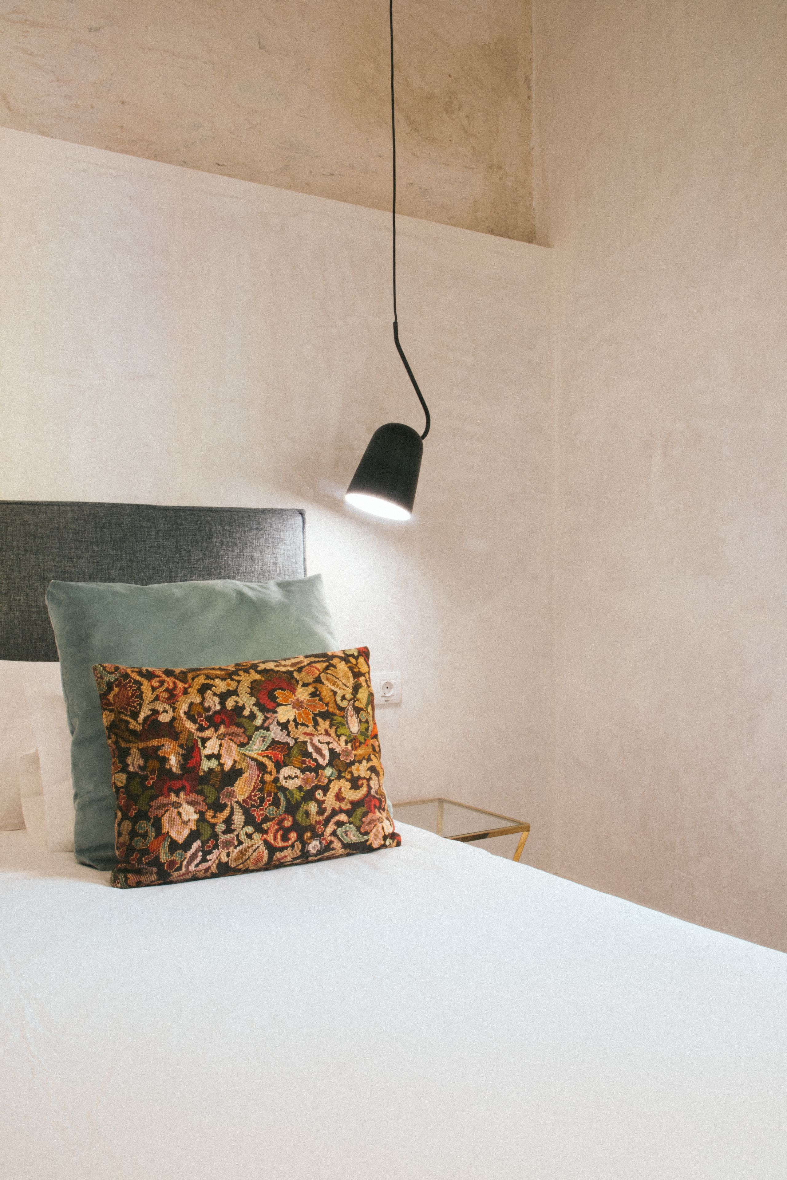 A bed with a white sheet, a green pillow, and a floral pillow. There is a black lamp hanging above the bed.