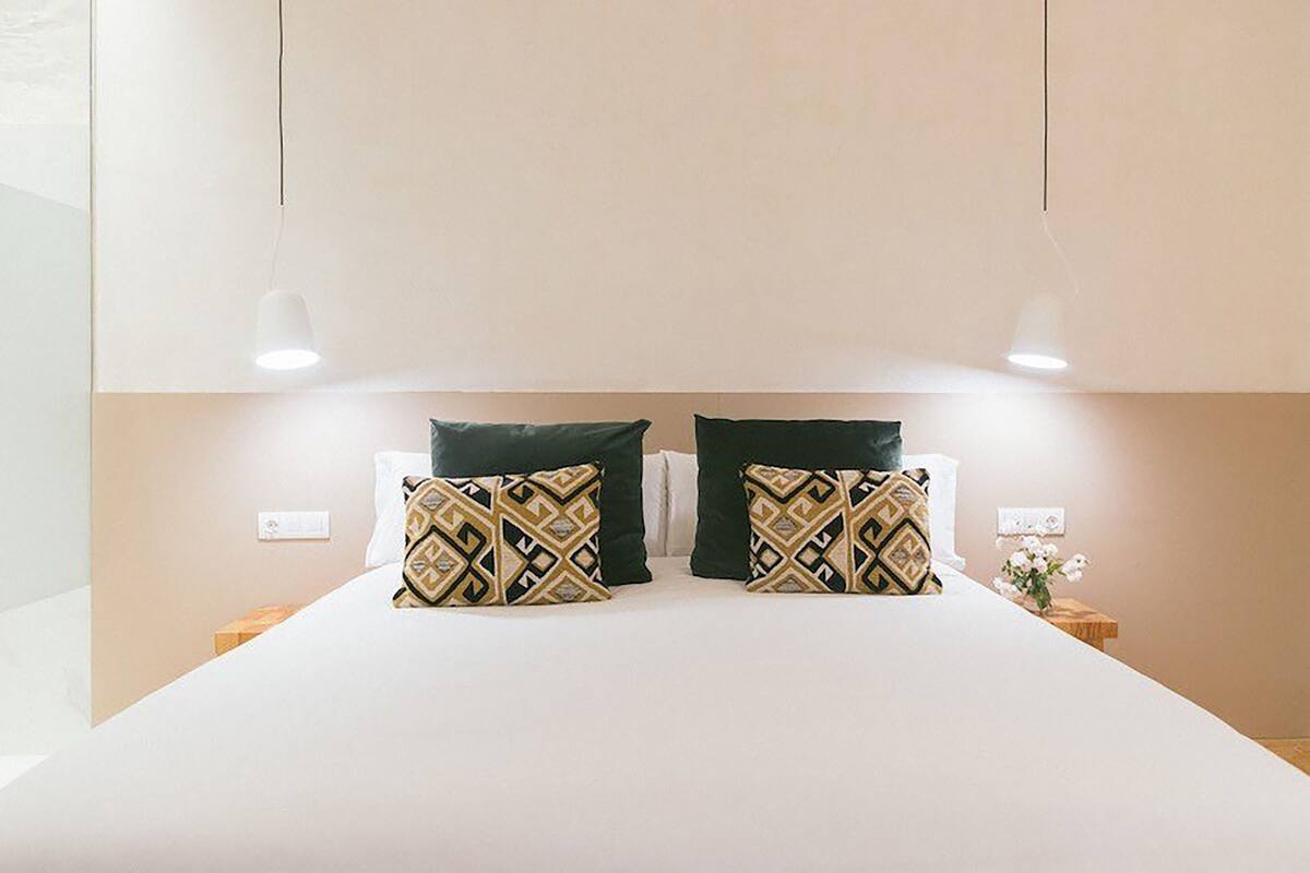 A neatly made bed with two decorative pillows, a bedside table with a vase of flowers, and a wall-mounted light fixture.