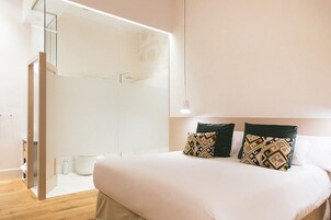 Una habitación de hotel moderna con una cama grande, dos almohadas estampadas, un box de ducha de vidrio y piso de madera.