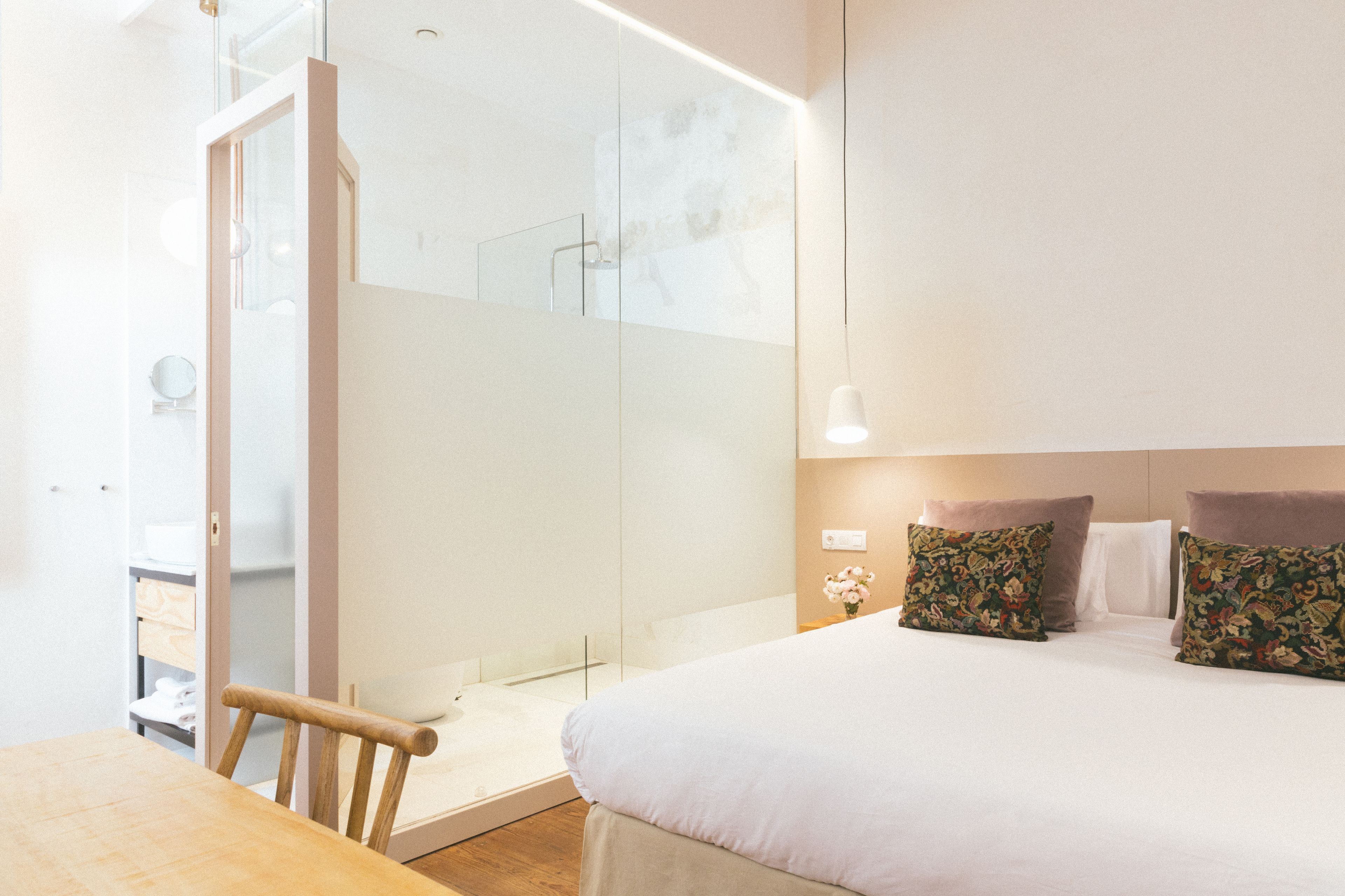 Un dormitorio moderno con una cama grande, una silla de madera, cabina de ducha de vidrio y una mesita.