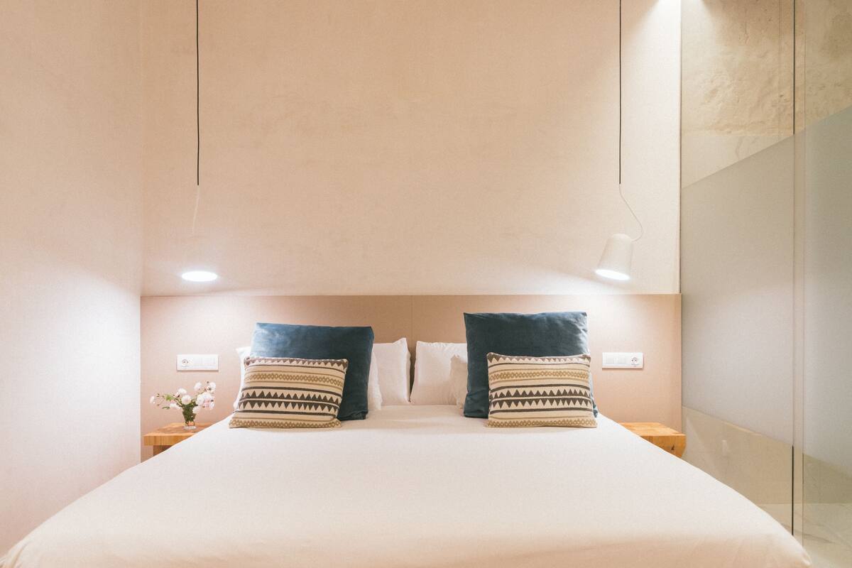A neatly made bed with two patterned pillows, a bedside table with a vase of flowers, and a wall-mounted light fixture.