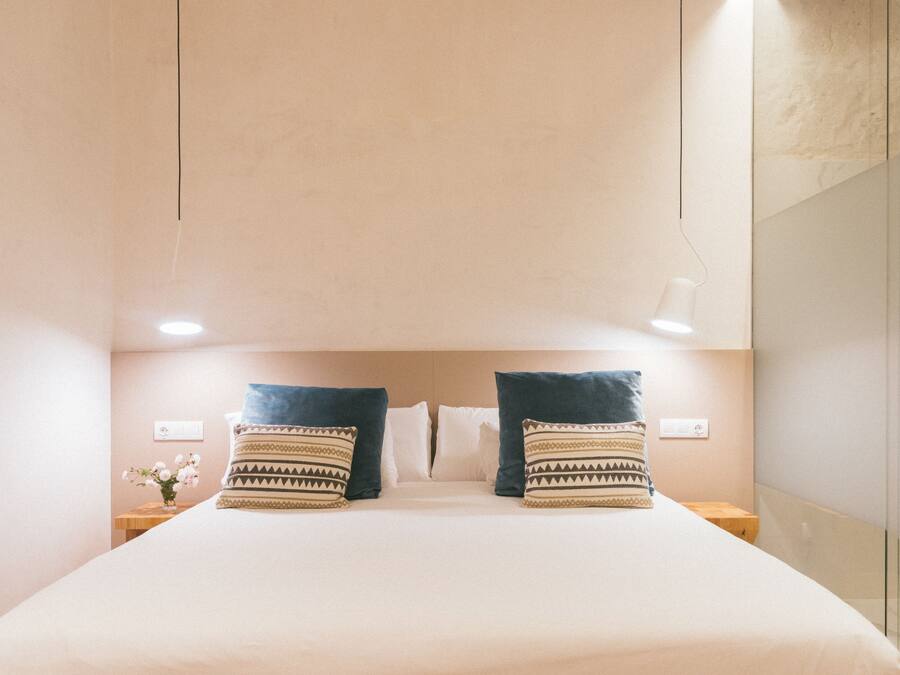 A neatly made bed with two patterned pillows, a bedside table with a vase of flowers, and a wall-mounted light fixture.