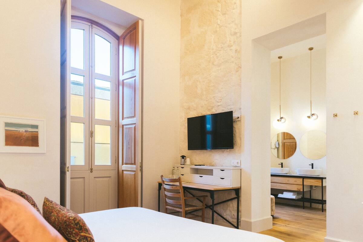 A bedroom with a bed, a desk, and a TV. There is a bathroom with a sink and mirror.