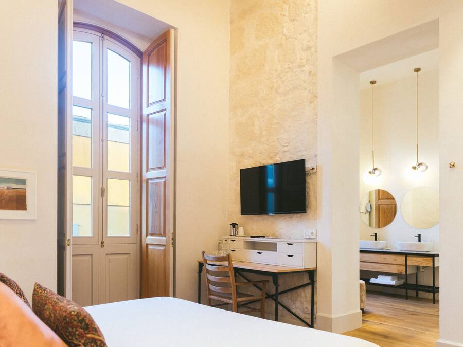 Un dormitorio con cama, escritorio y televisor. Cuenta con un baño con lavamanos y espejo.
