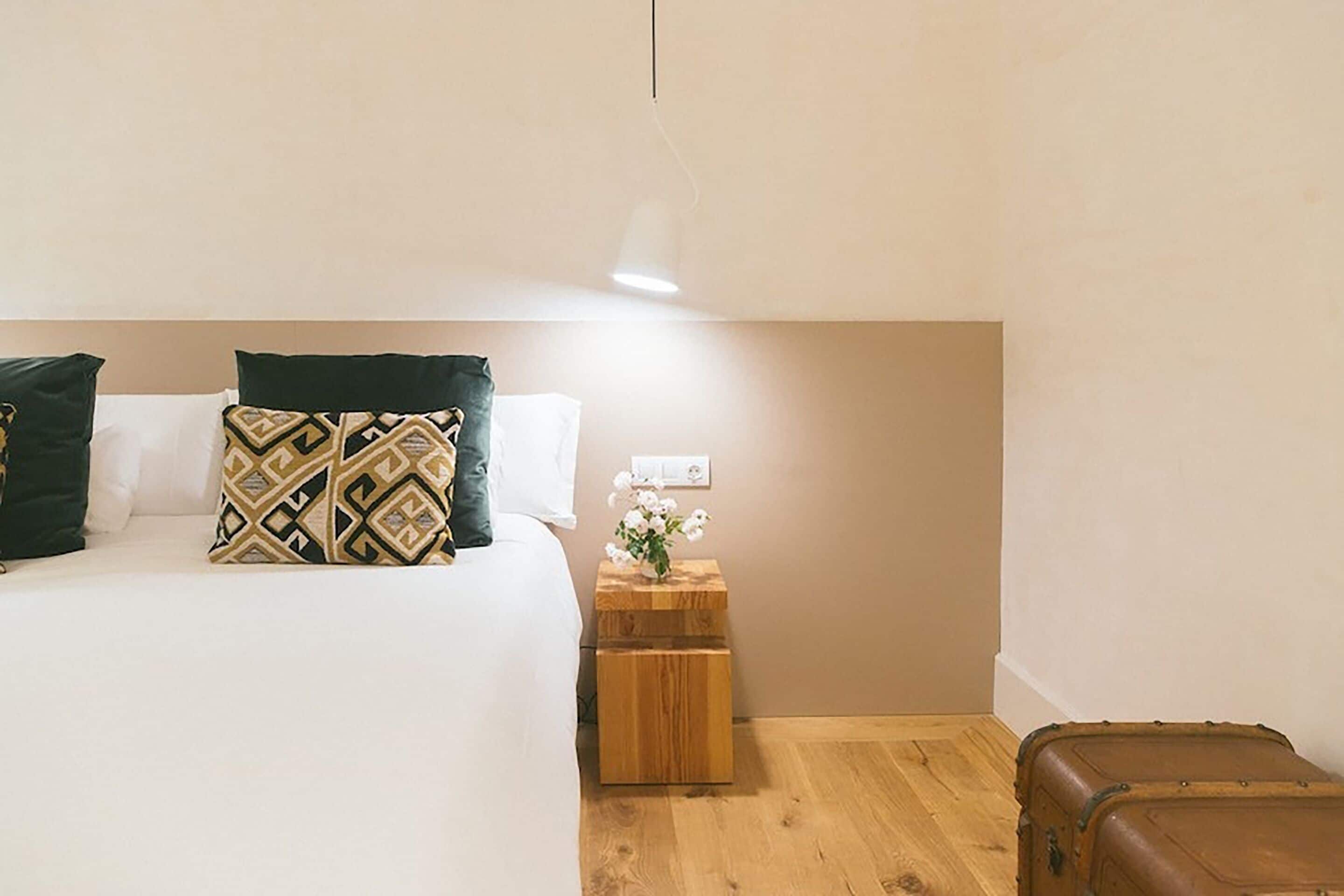 A bedroom with a bed, pillows, a nightstand with a vase of flowers, and a suitcase.