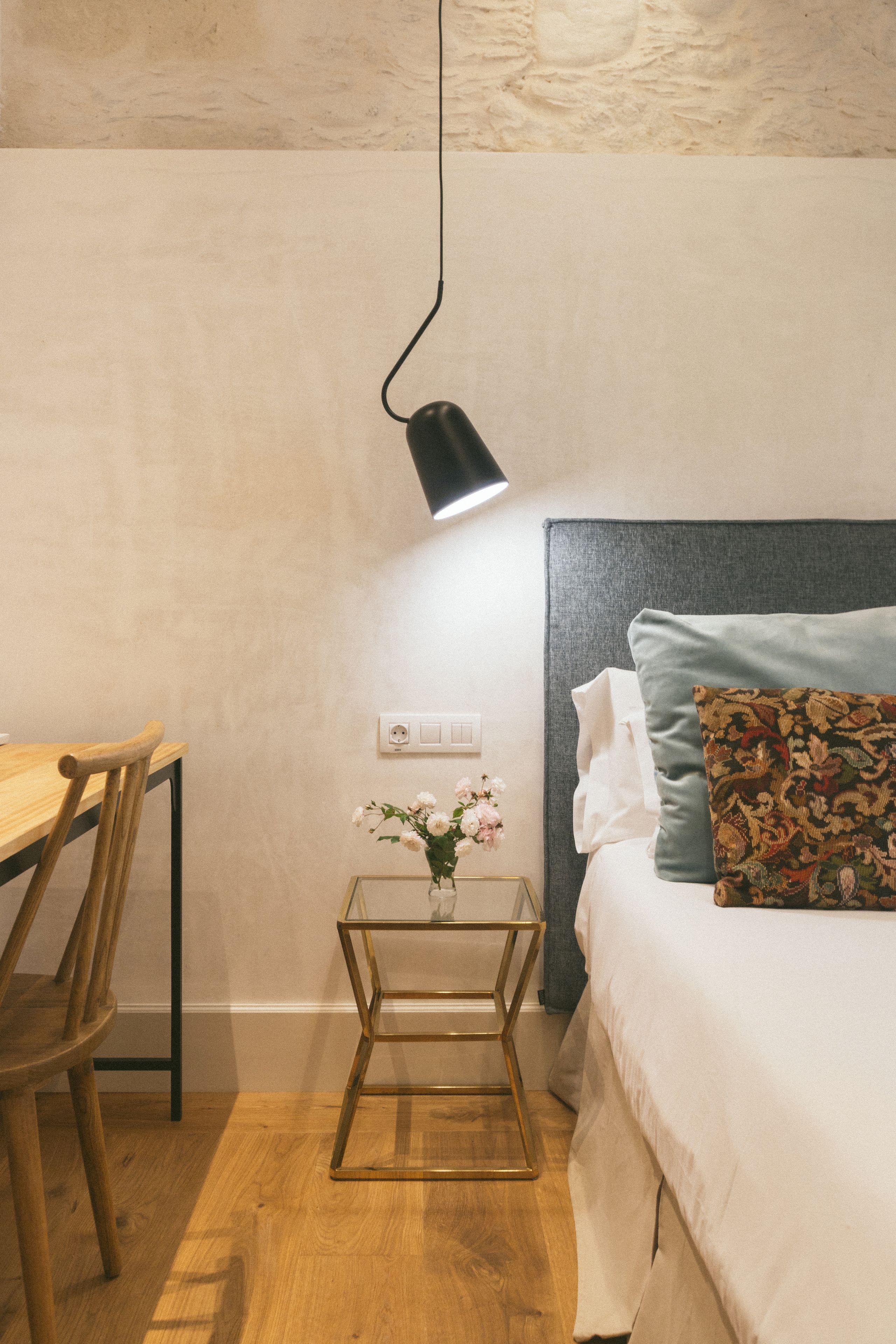 Un dormitorio con una silla de madera, una mesita de noche con un jarrón de flores, una cama con un cojín estampado e iluminación en la pared.