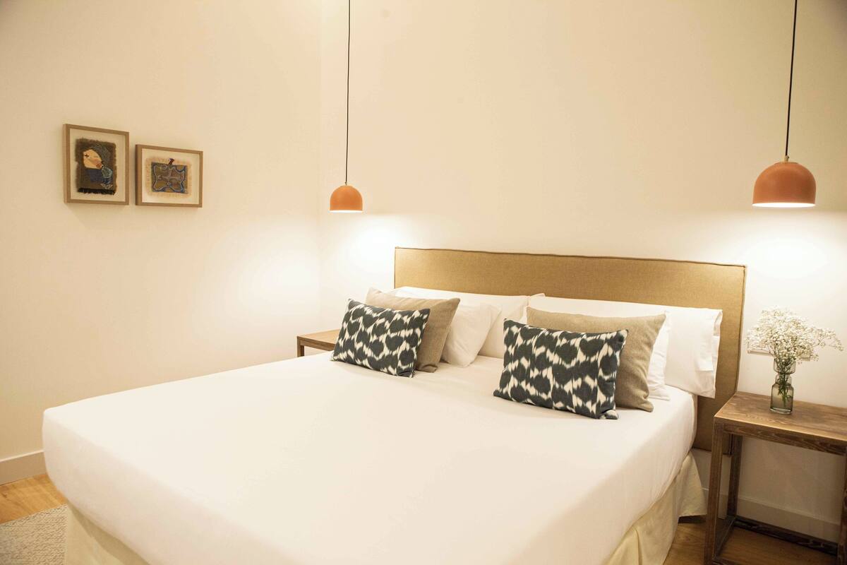 A neatly made bed with white linens and patterned pillows, a wooden headboard, and two pendant lights.