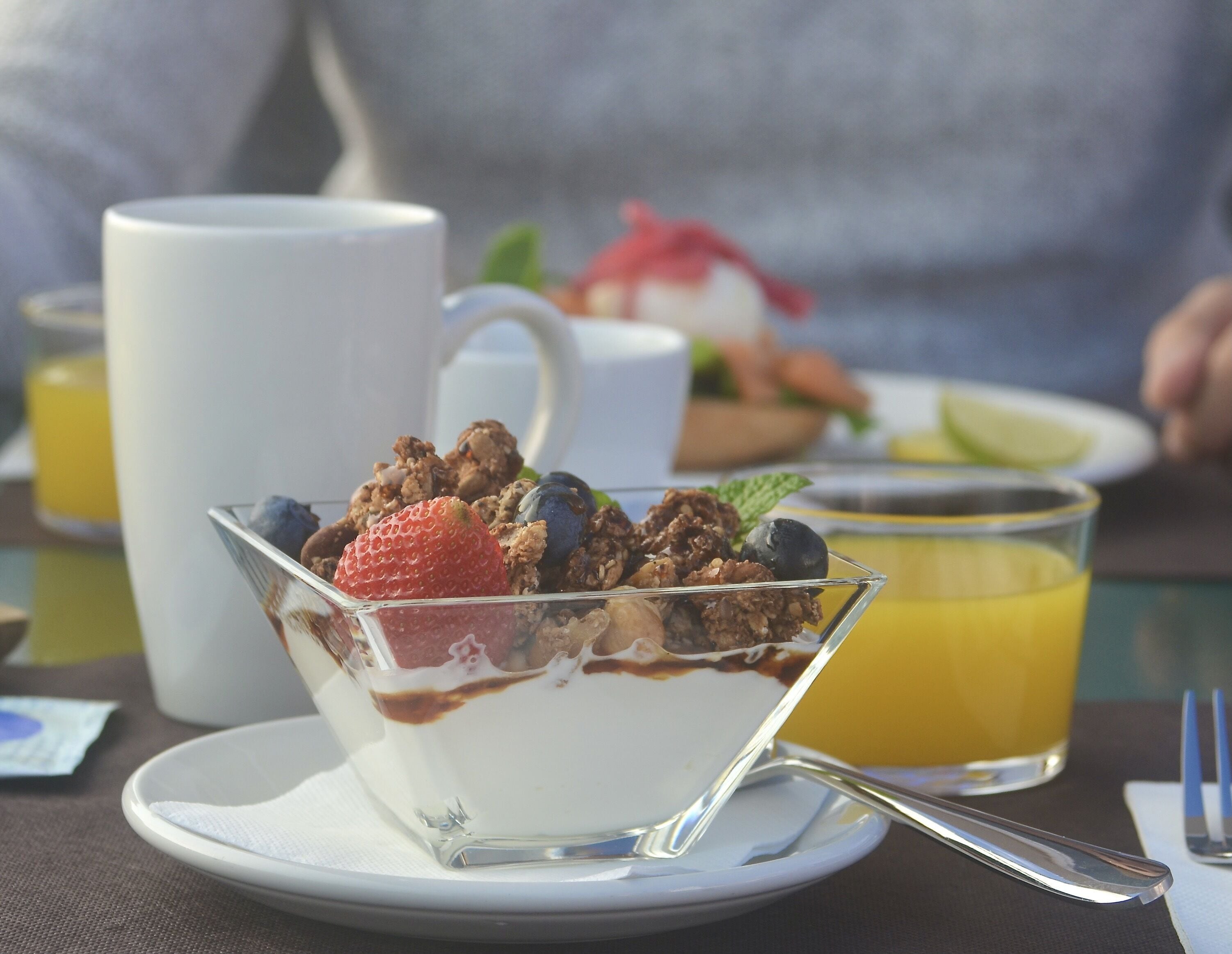 Un postre en un bol de cristal sobre un plato, con una cuchara y un vaso de jugo de naranja al fondo.
