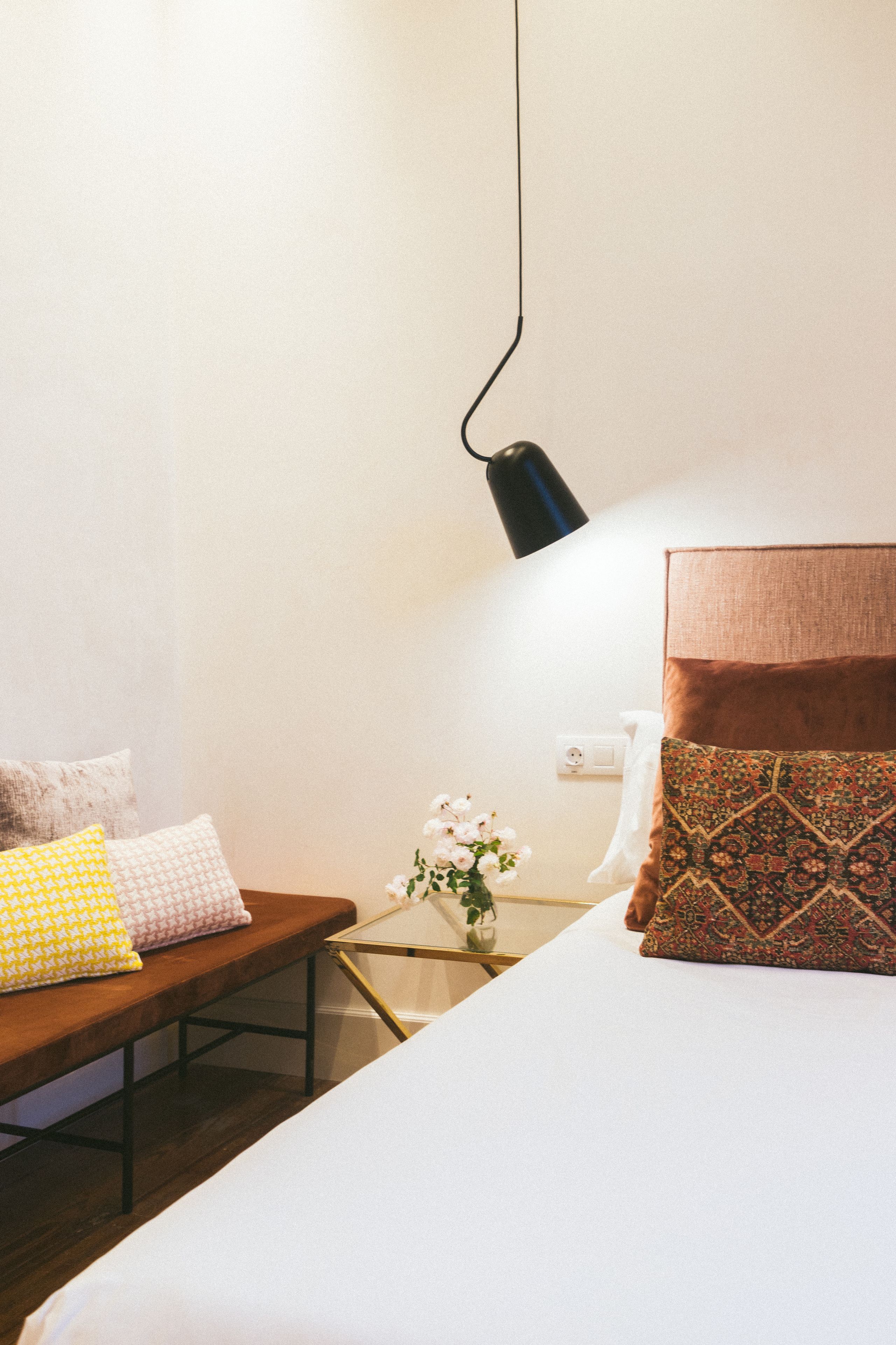 Un dormitorio ordenado con cama, mesita de noche, lámpara y un pequeño jarrón con flores.