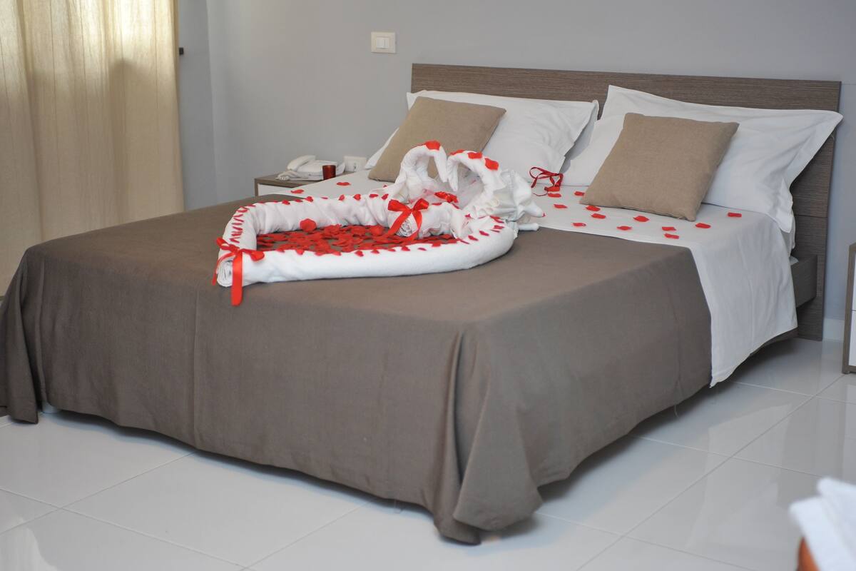 A neatly made bed with a brown bedspread, white pillows, and a decorative arrangement of red rose petals and a white cloth with red ribbons.