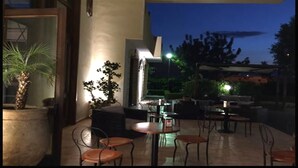 Un área de estar al aire libre con mesas y sillas, rodeada de plantas en macetas y vista al cielo nocturno.