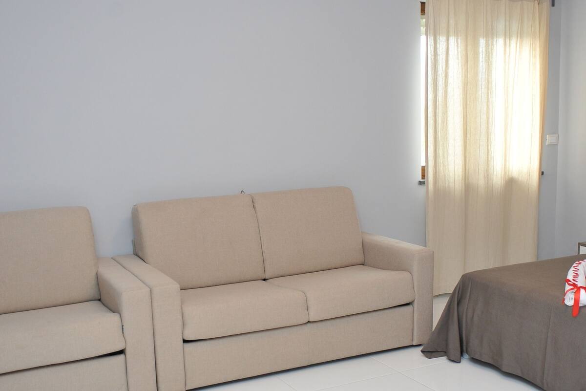 A room with a beige sofa, a bed with a brown cover, and a window with beige curtains.