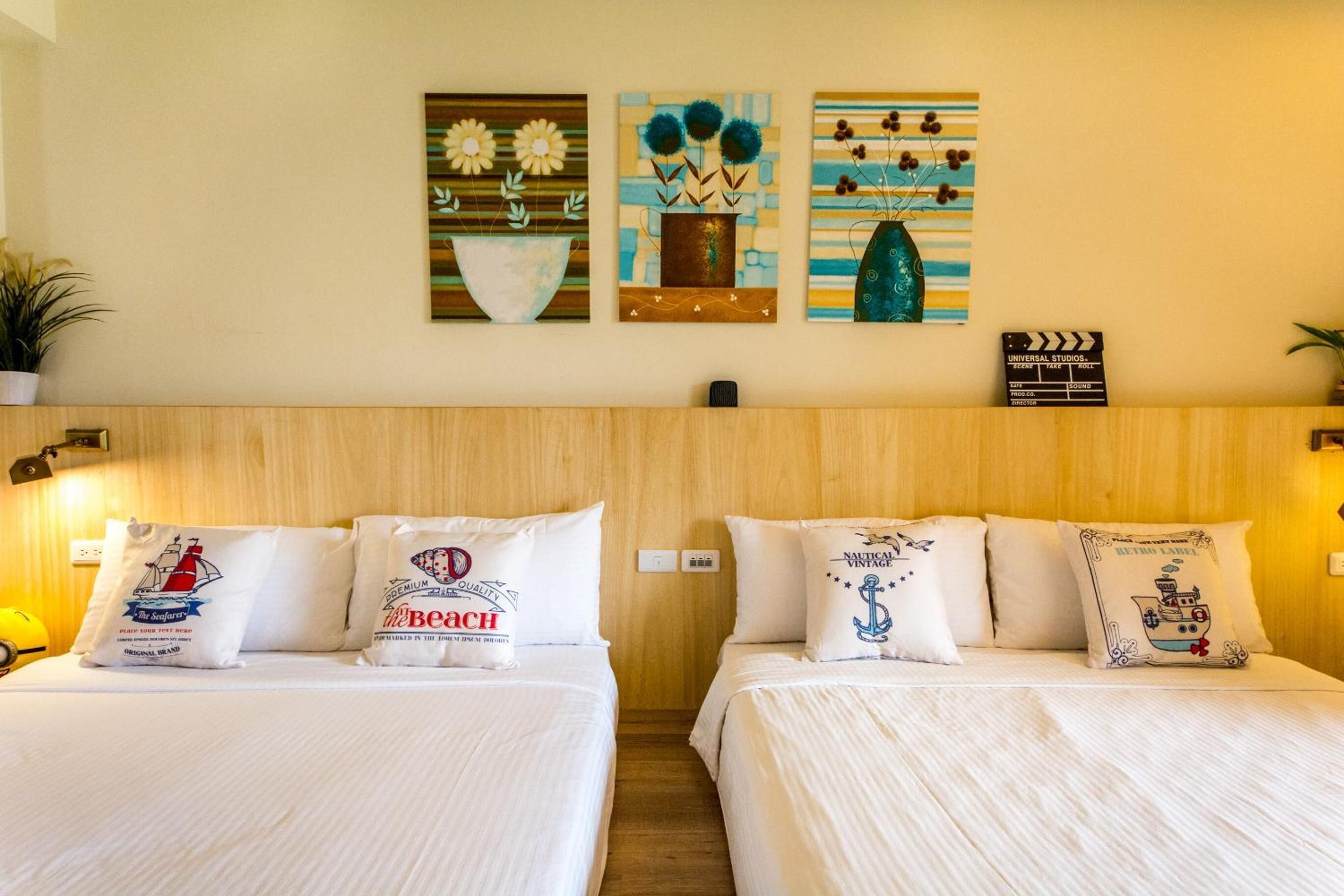 A hotel room with two beds, each with white bedding and decorative pillows. There are wall-mounted artworks above the beds, and a clapperboard sign on the wall.