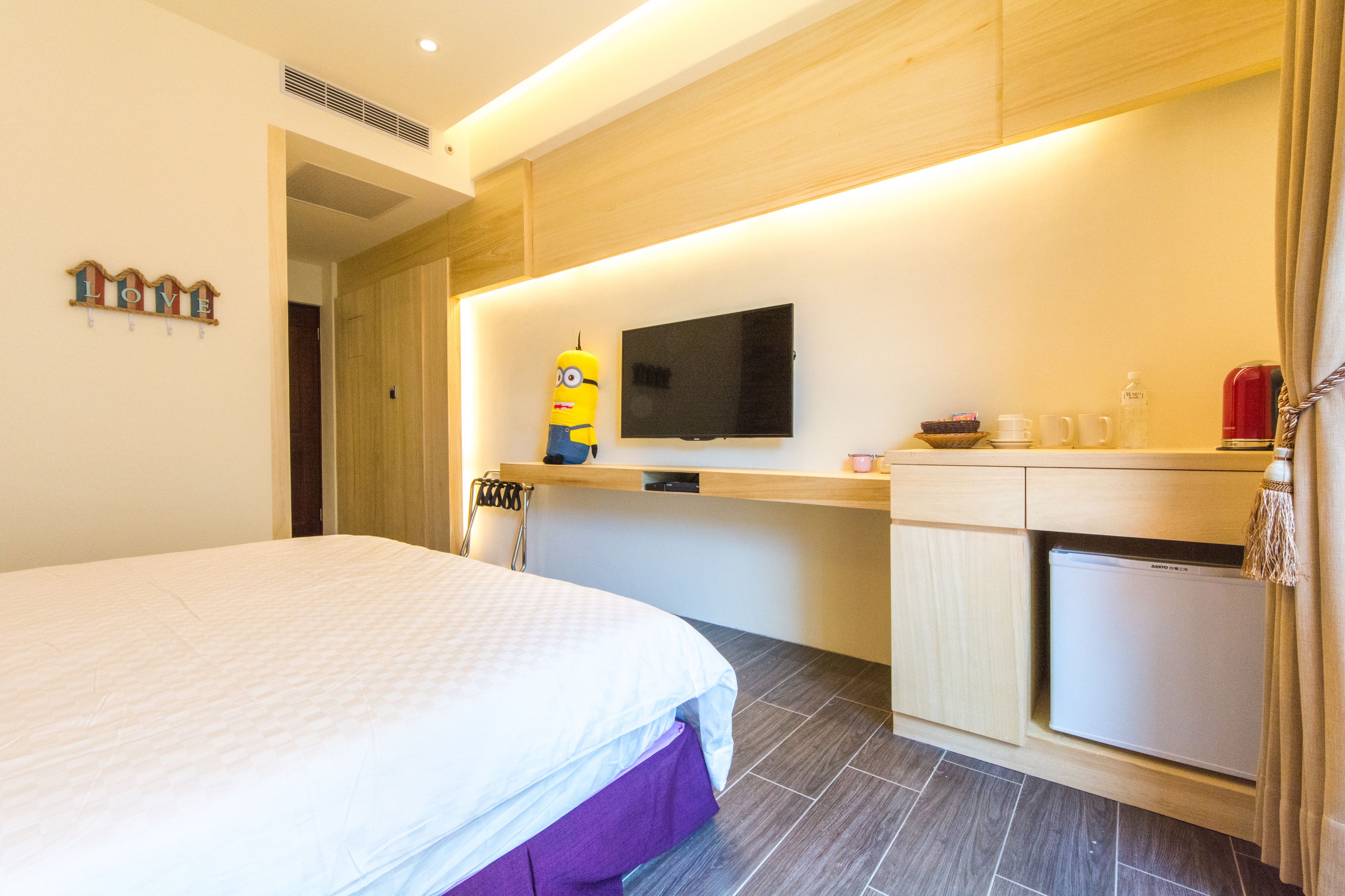 A hotel room with a bed, a small kitchenette, a television, and a Minion plush toy.