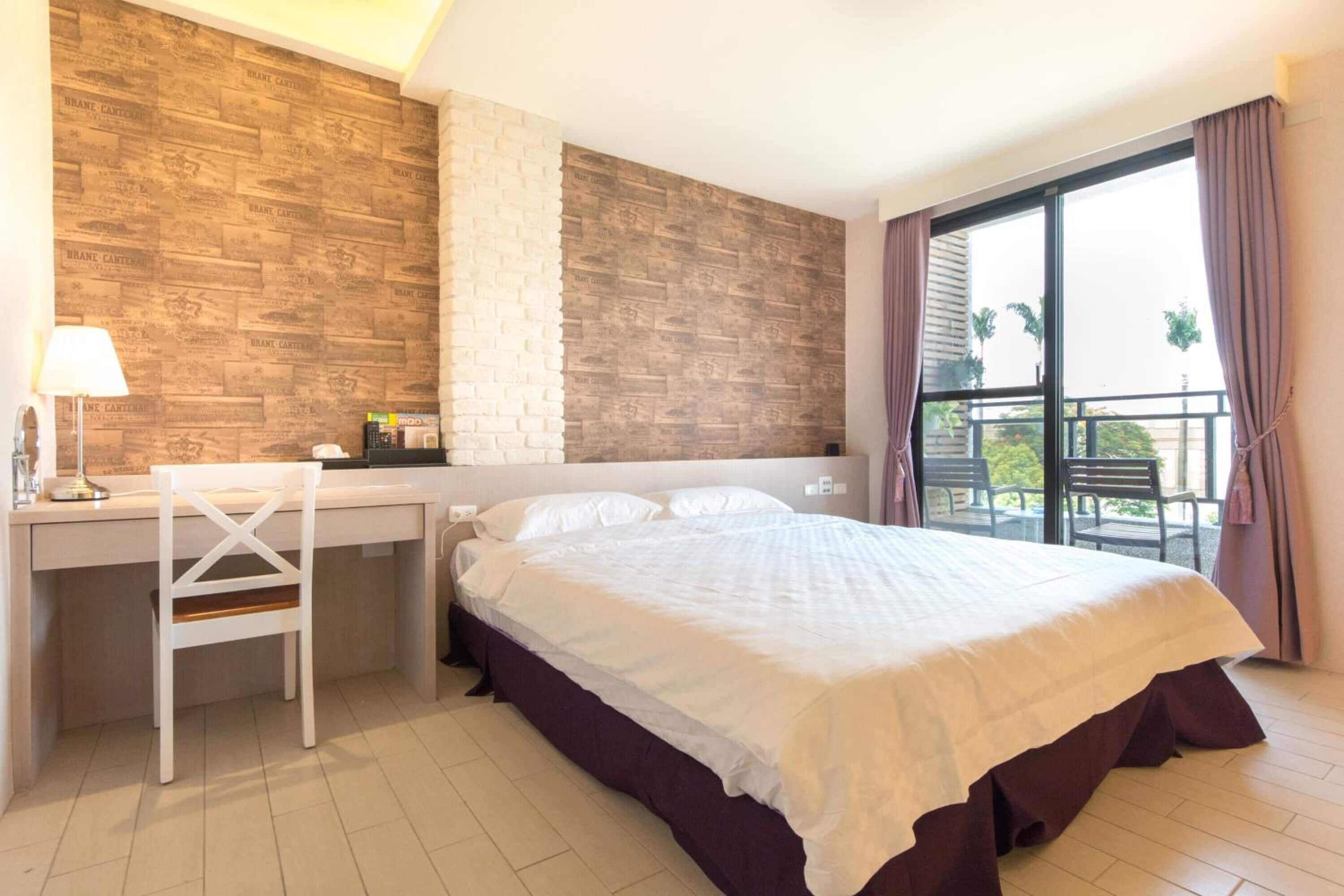 A bedroom with a bed, desk, chair, and a balcony with curtains.
