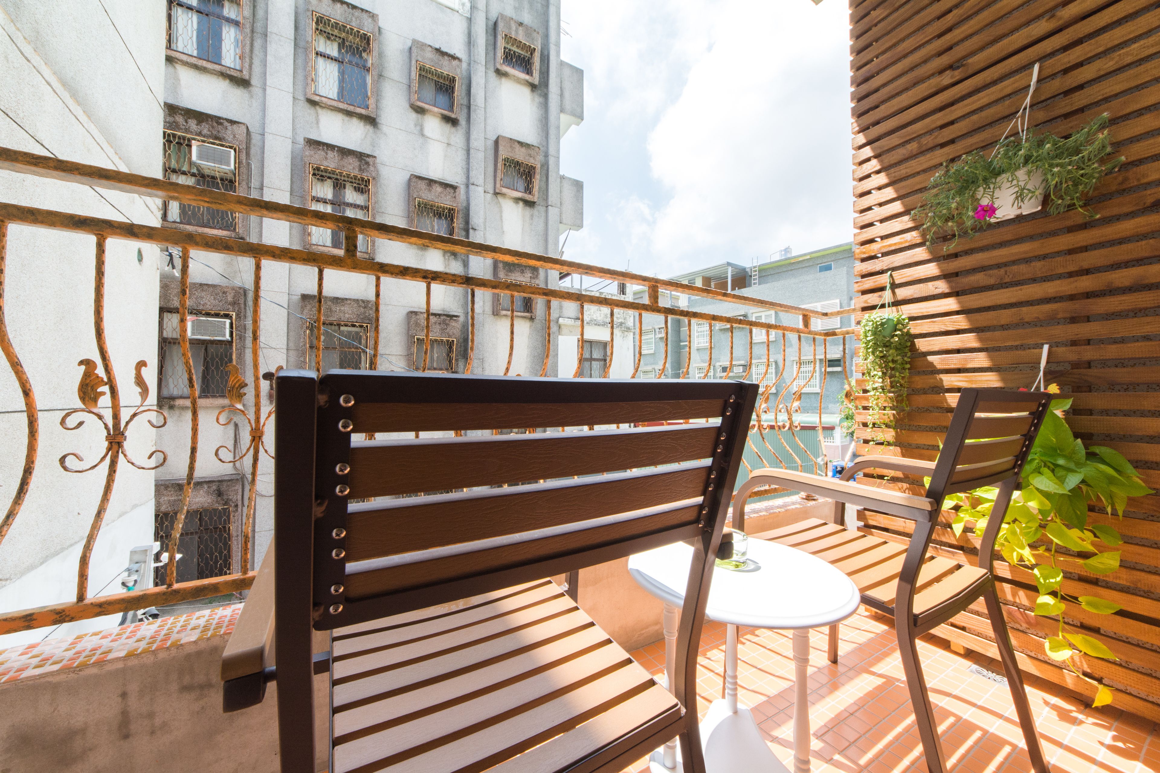 A balcony with a wooden chair and a small table, overlooking a building with multiple windows and a metal railing.