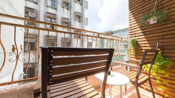 A balcony with a wooden chair and a small table, overlooking a building with multiple windows and a metal railing.