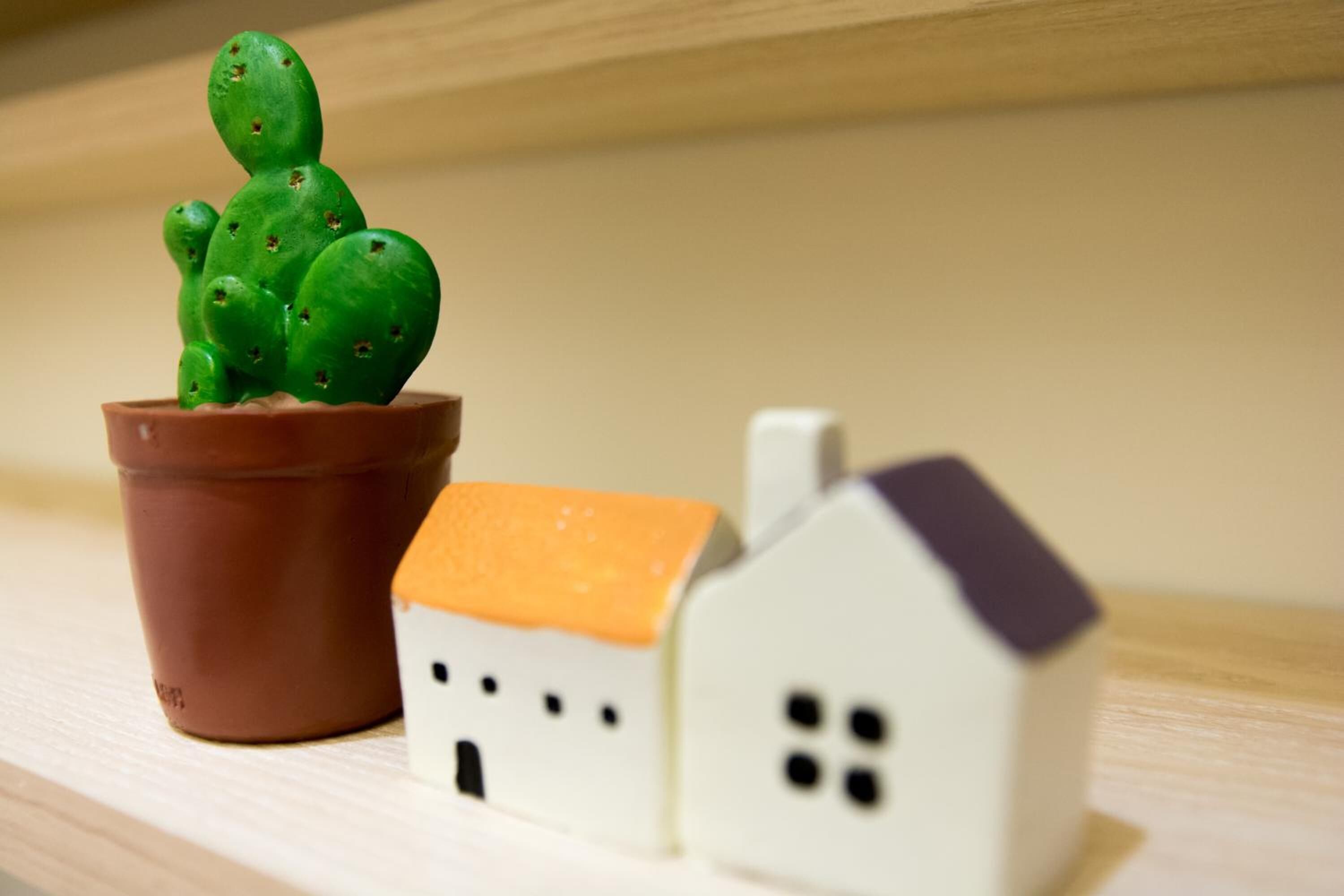 Ein kleiner Kaktus in einem braunen Topf neben einem Miniatur-Weißen Haus mit brauner Dacheindeckung.
