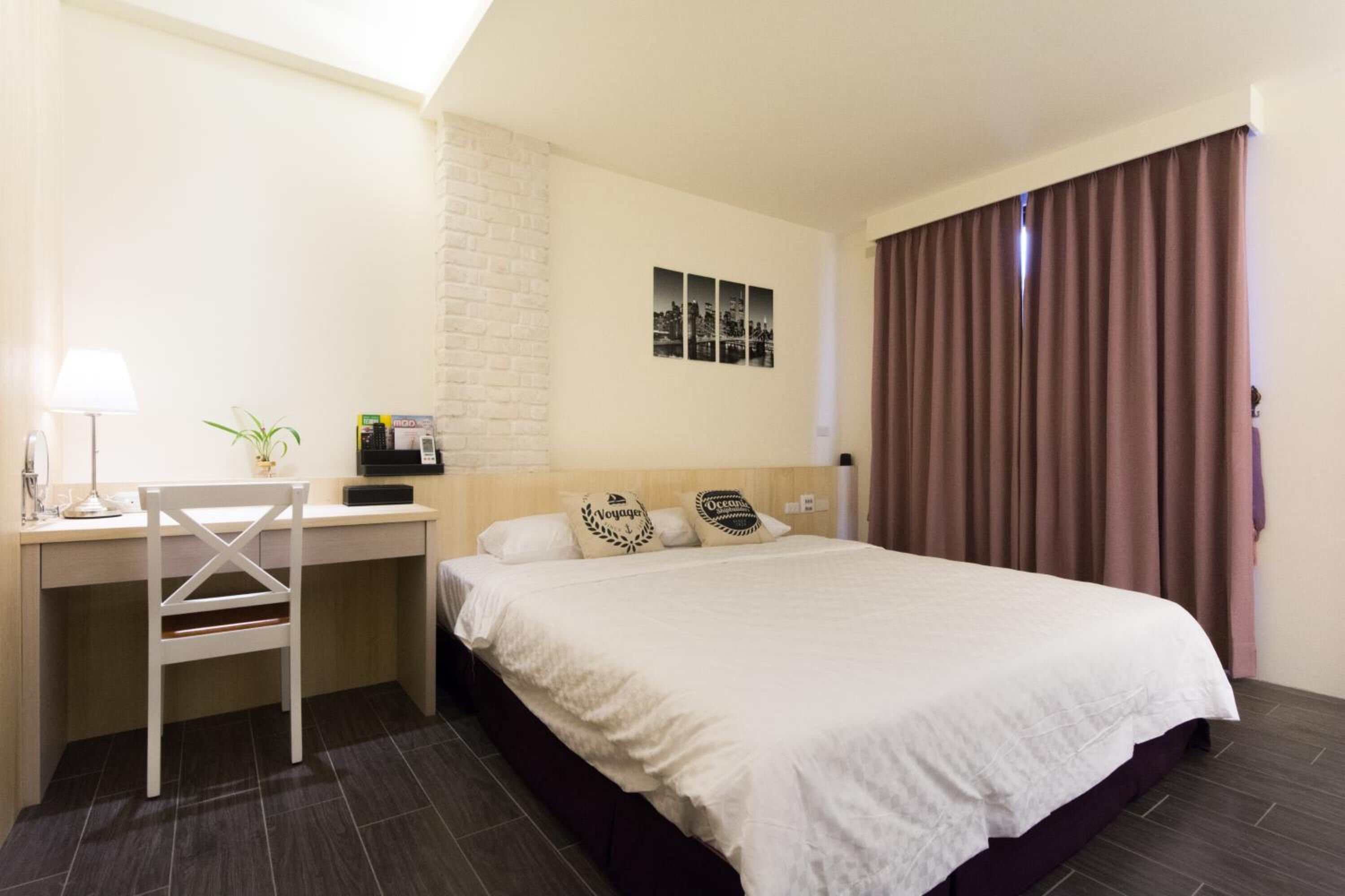 A hotel room with a bed, a desk with a chair, a lamp, a plant, and a cityscape picture on the wall.