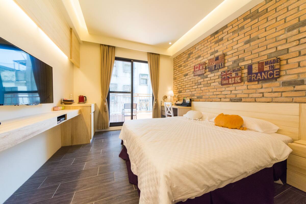 Una habitación de hotel con una cama grande, un escritorio, un televisor y una pared de ladrillo con carteles.