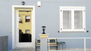 Una entrada de edificio con puerta, ventana con contraventanas, banco, mesa y farol.