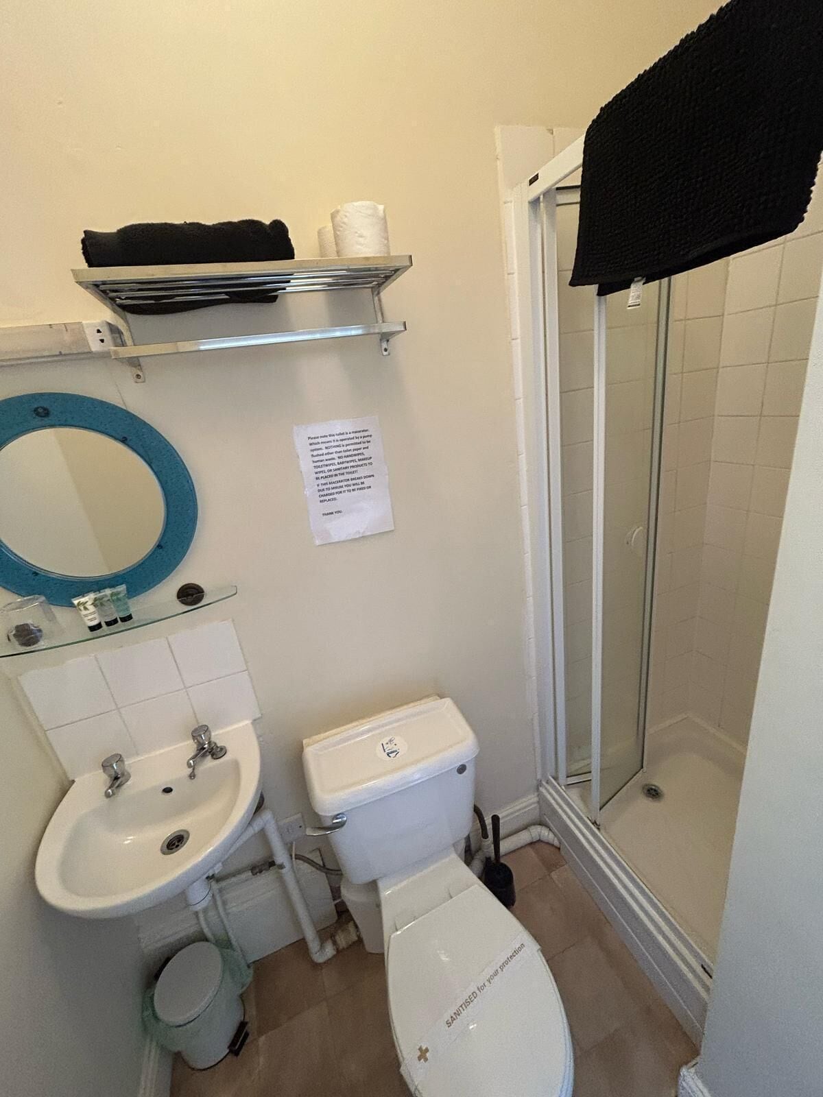 Een badkamer met een wastafel, toilet en douche. Er is een spiegel, een plank met handdoeken, en een bordje aan de muur.