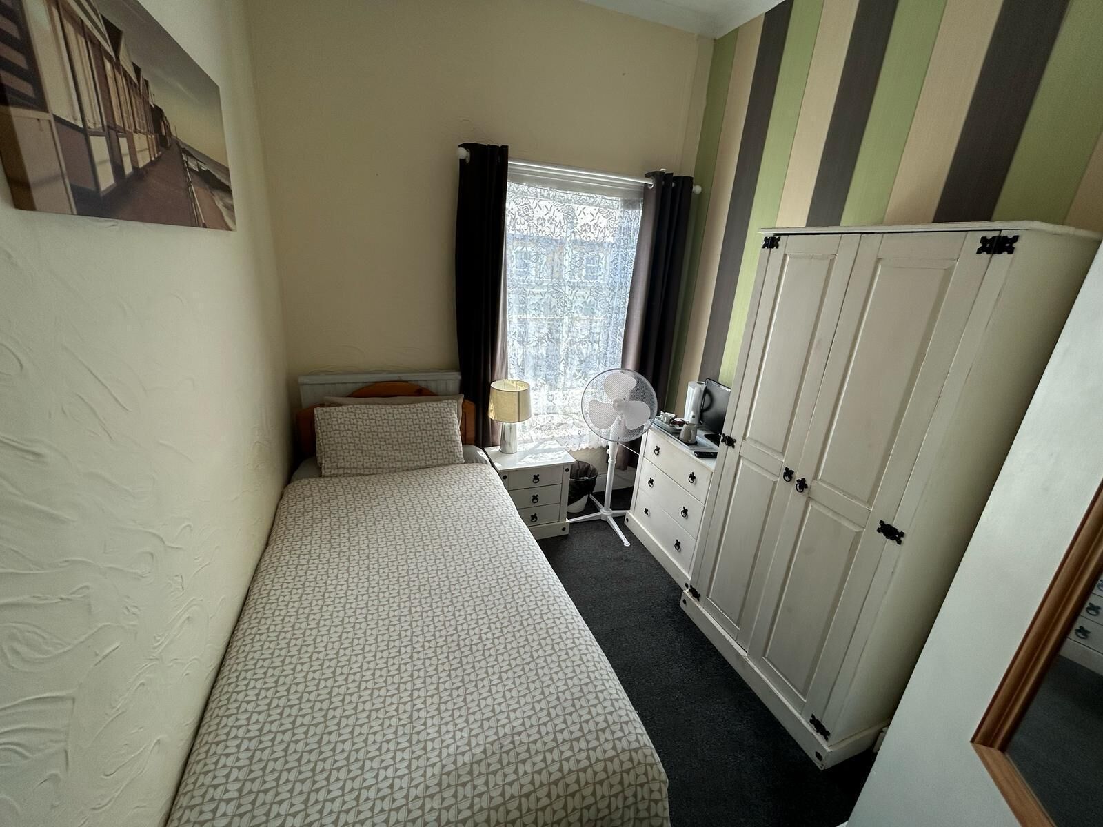 A small, neatly arranged bedroom with a single bed, a bedside table with a lamp, a fan, and a wardrobe.
