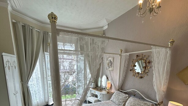 A bedroom with a four-poster bed, a chandelier, a bedside table with a lamp, and a window with curtains. - The little emily - Room only (Great Yarmouth)