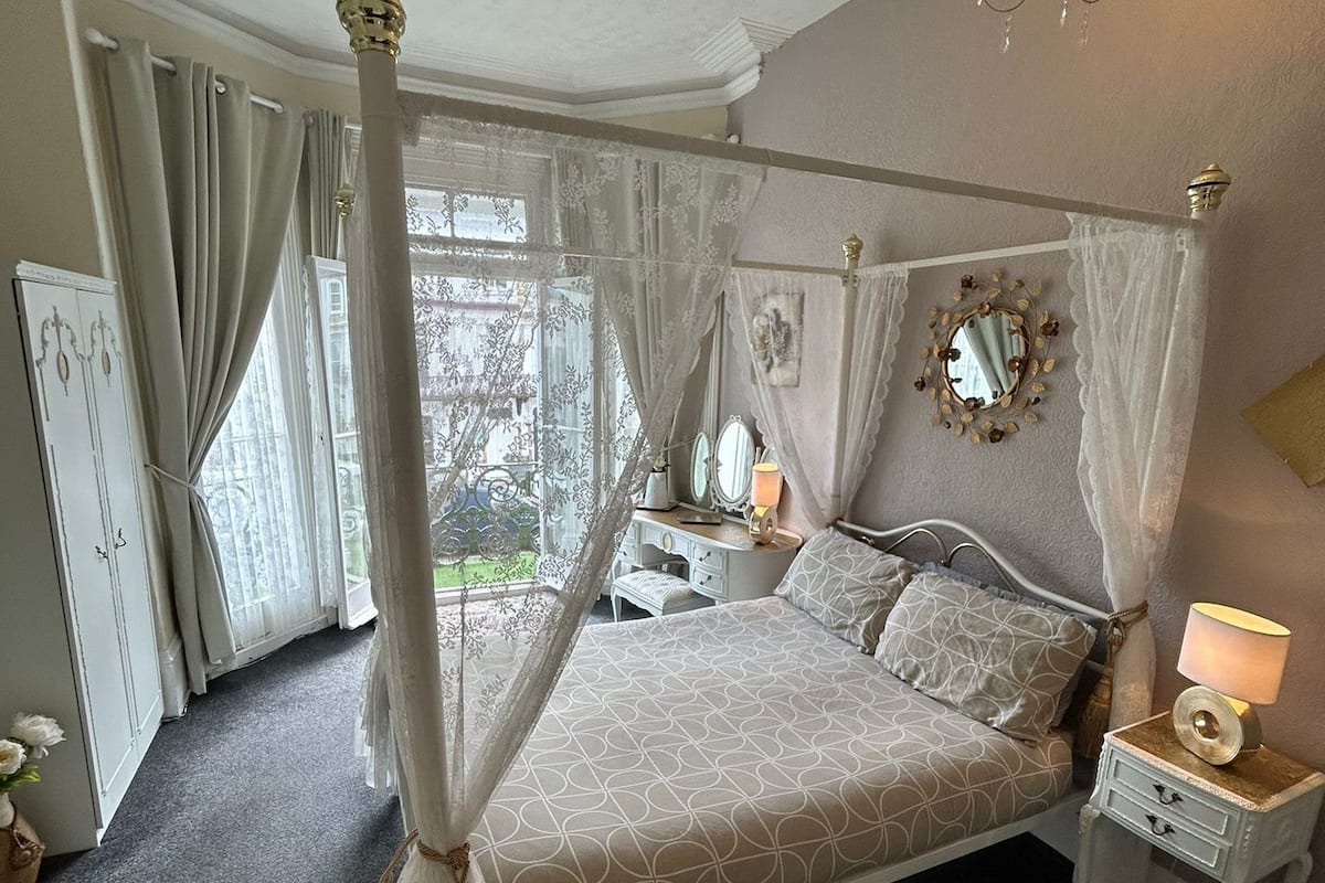 Un dormitorio con cama con dosel, candelabro, mesita de noche con lámpara y ventana con cortinas.