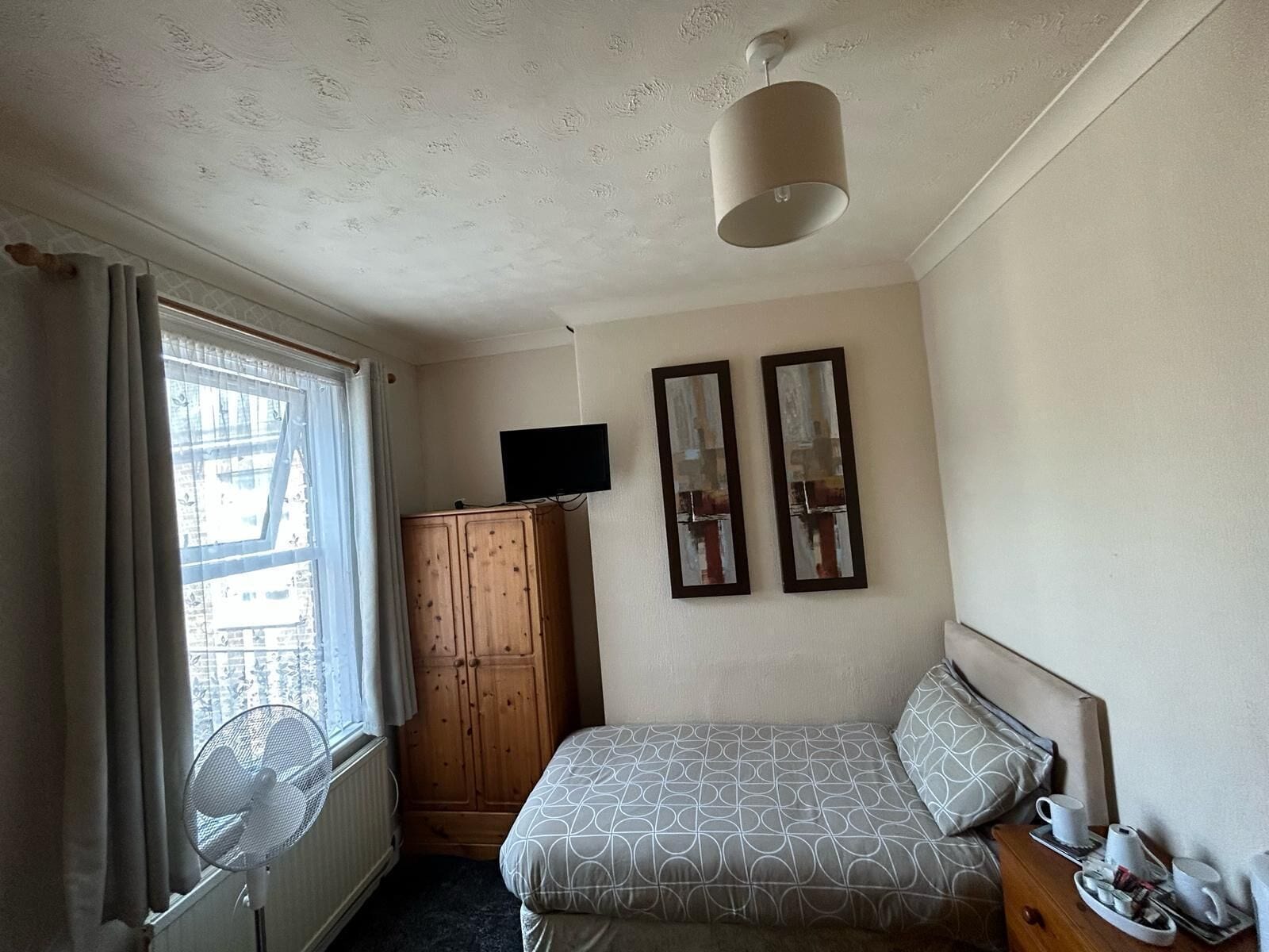 Een hotelkamer met een bed, een houten garderobe, een flatscreen-tv, twee ingelijste foto's, een plafondventilator en een raam met gordijnen.