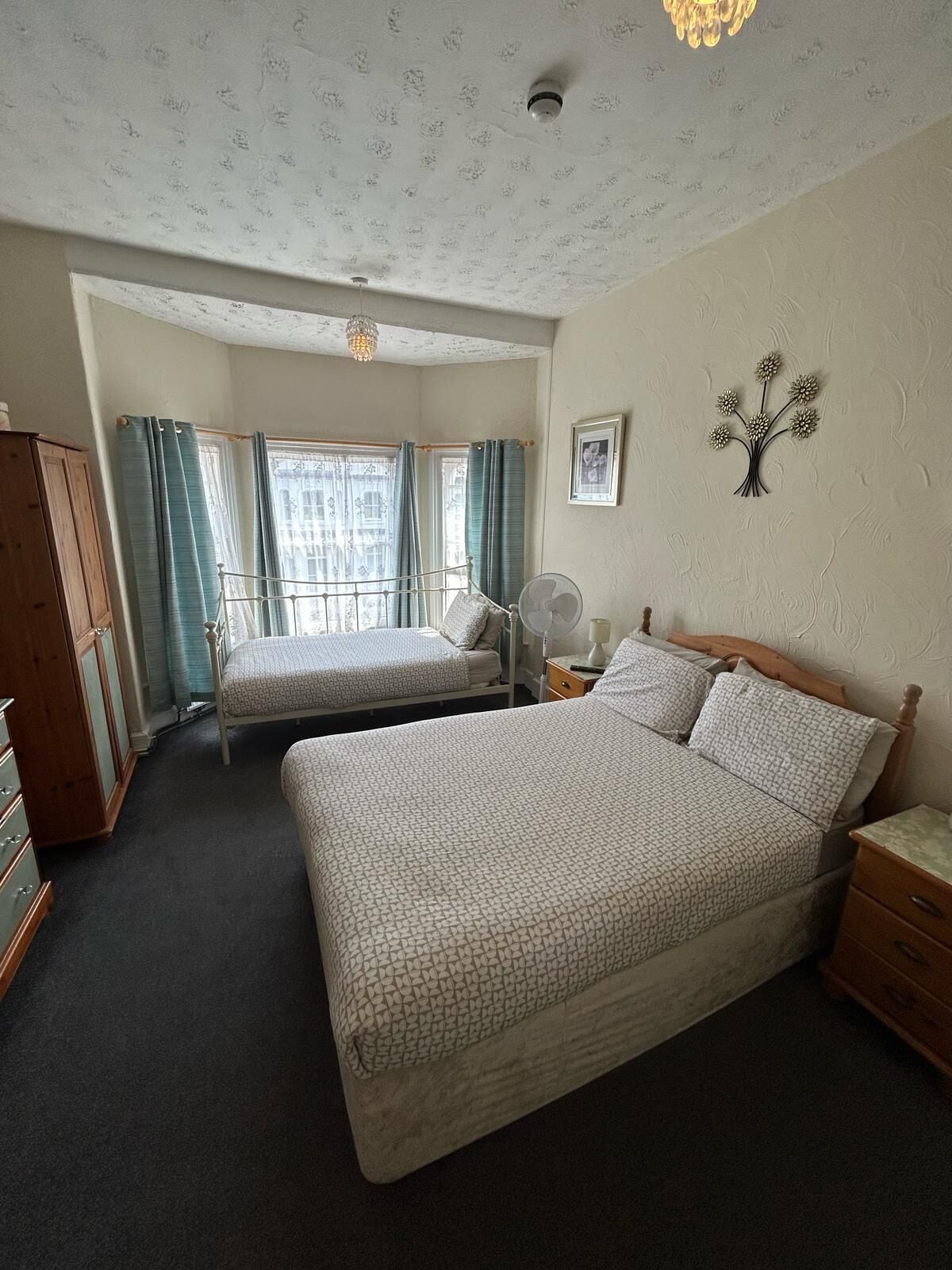 A bedroom with two beds, a wooden wardrobe, a bedside table with a lamp, and a wall-mounted fan.