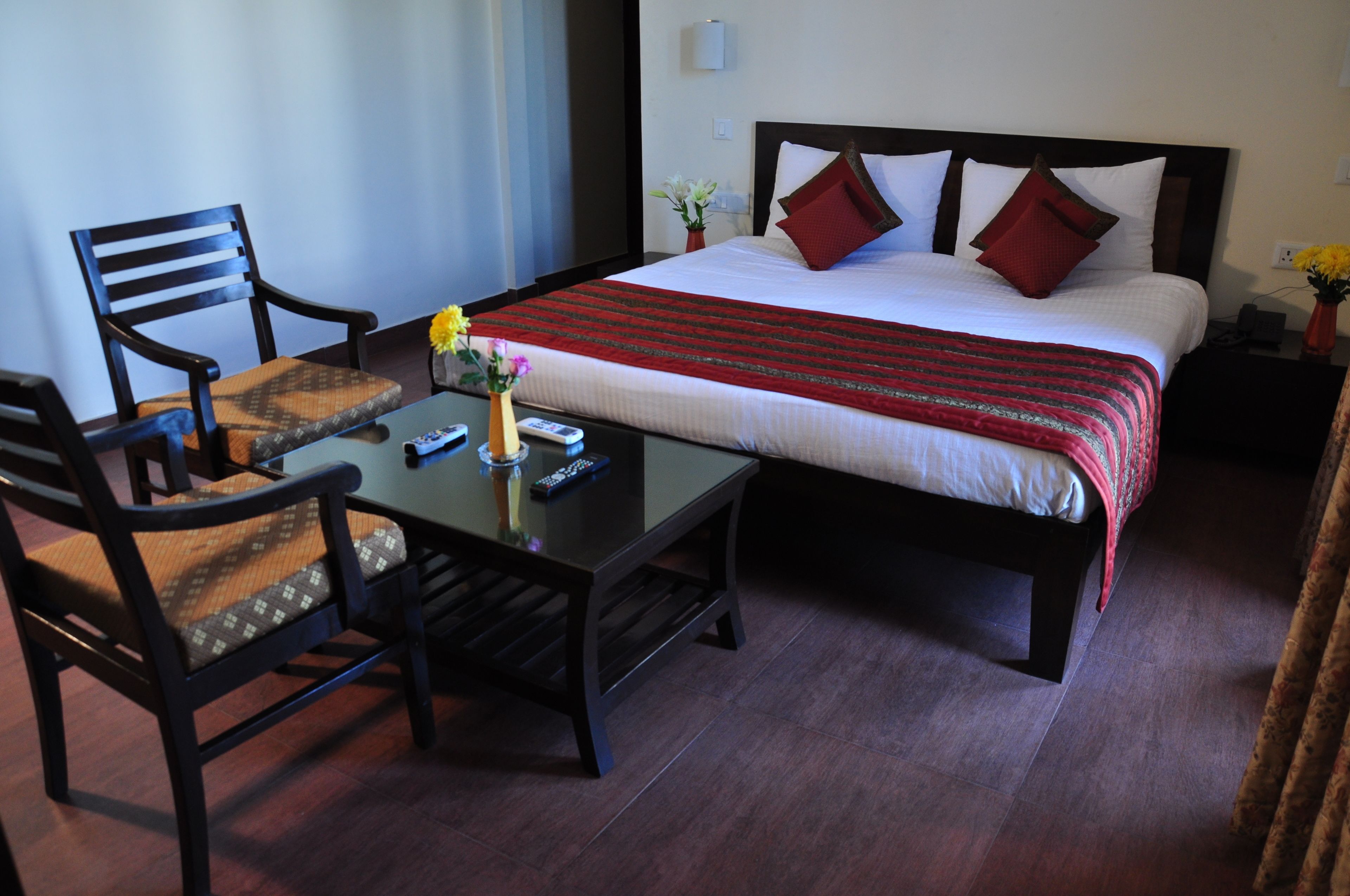 Habitación de hotel con cama, dos sillas, una mesita con un jarrón y controles remotos, y una mesita de noche con lámpara y flores.
