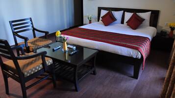 Habitación de hotel con cama, dos sillas, una mesita con un jarrón y controles remotos, y una mesita de noche con lámpara y flores.