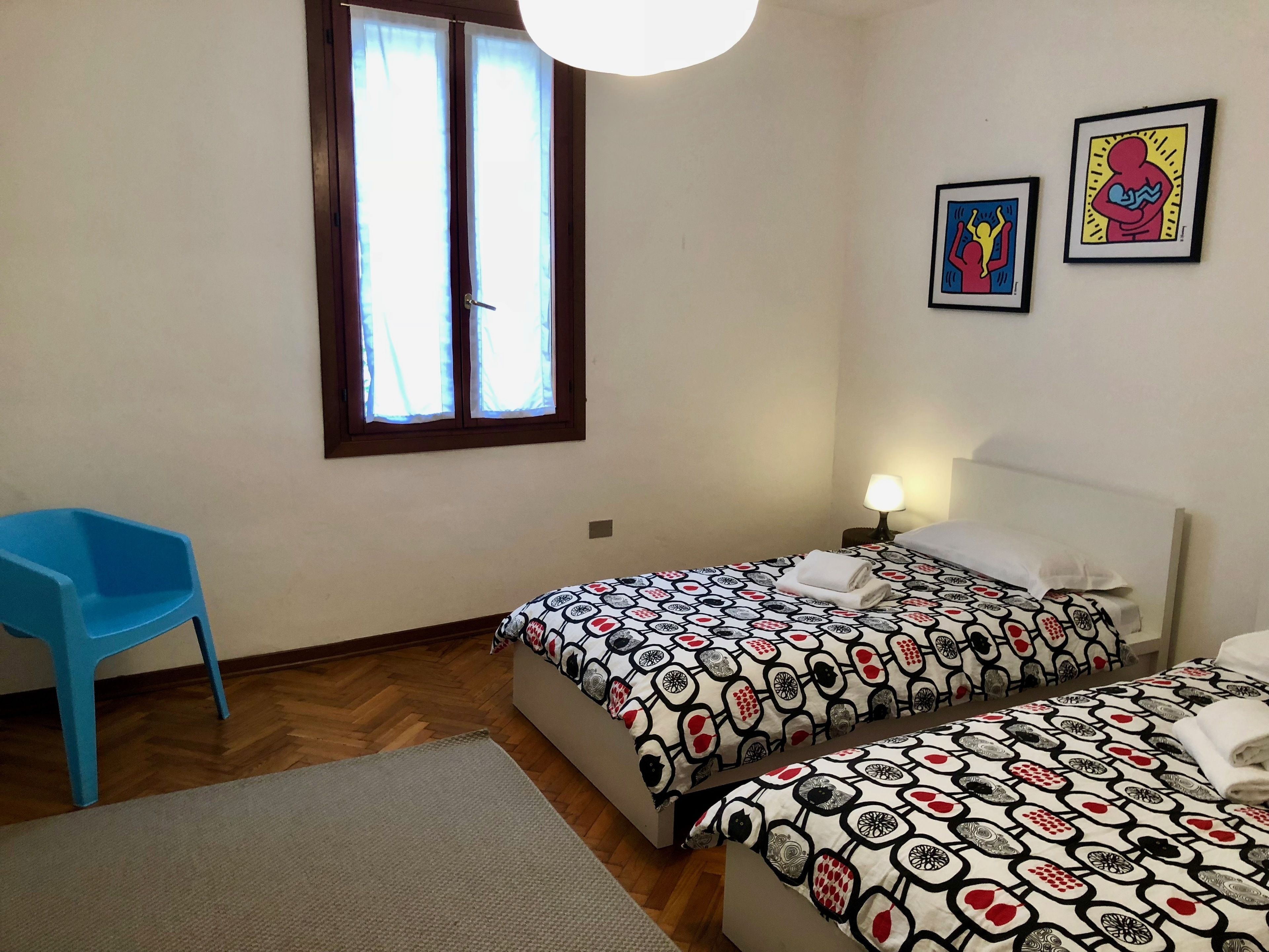 Une chambre d’hôtel avec deux lits, une chaise bleue, une fenêtre avec des rideaux et deux tableaux encadrés accrochés au mur.