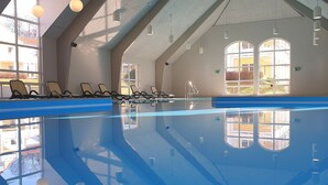 Piscina interior com paredes de vidro transparente, cadeiras de descanso ao lado e luz natural entrando por grandes janelas.