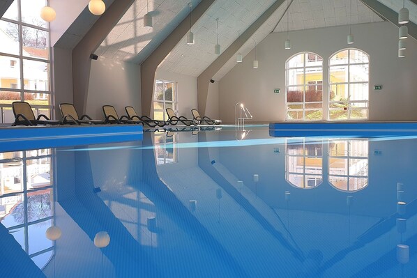 Une piscine intérieure aux parois en verre transparent, dotée de chaises longues le long des bords et baignée de lumière naturelle grâce à de grandes fenêtres.