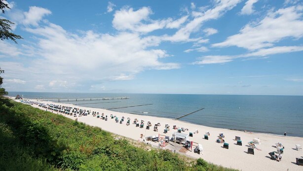 Uma praia com fileiras de cadeiras e guarda-sóis de praia, um píer que se estende para a água, e um céu claro.